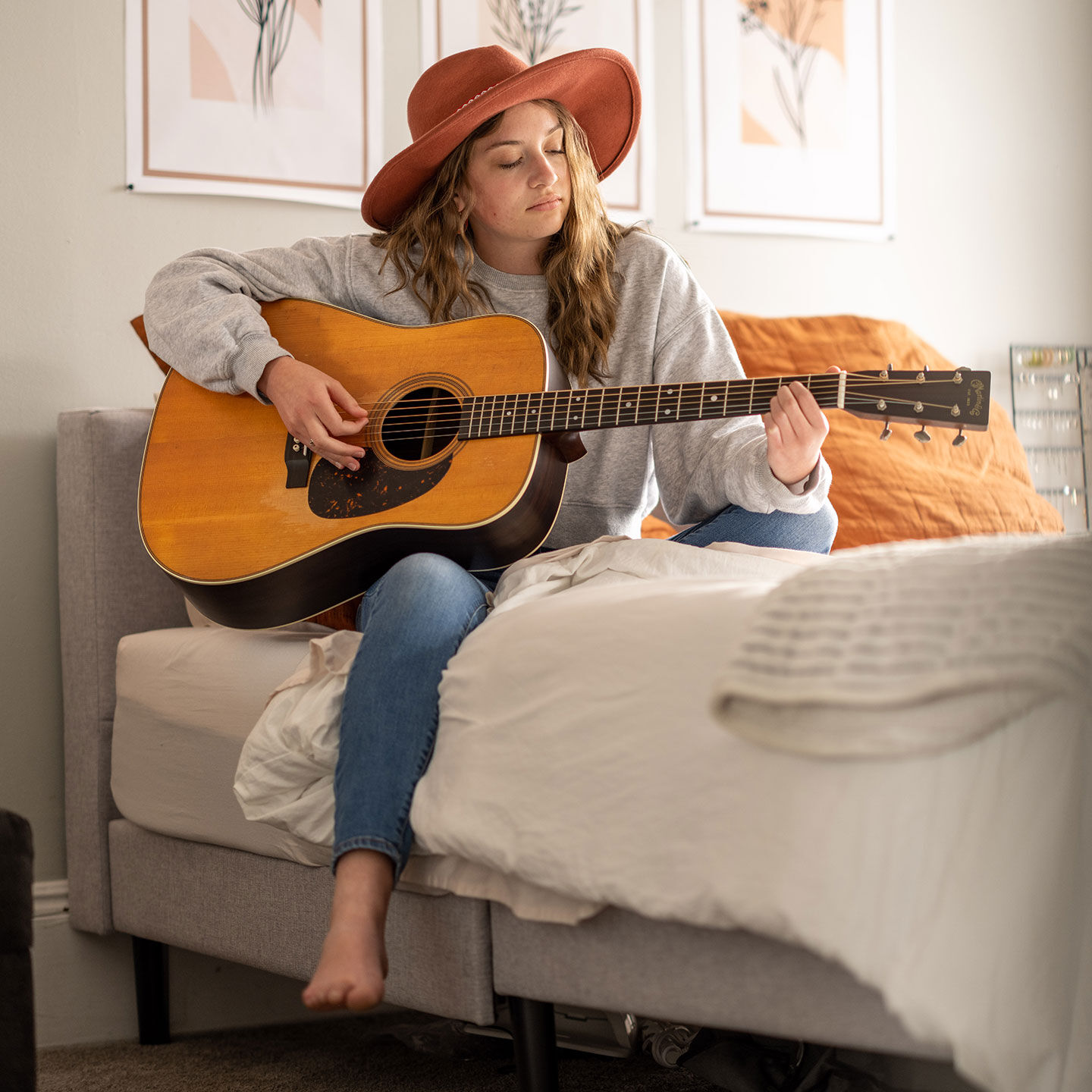 Young woman in a red brimmed hat sitting on a bed playing a Martin dreadnought.